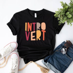 May include: Black t-shirt with the word "INTROVERT" printed in colourful block letters. The shirt is laid flat on a white surface with a pair of white trainers, a wooden bead necklace, a pair of denim shorts, and a pair of sunglasses.