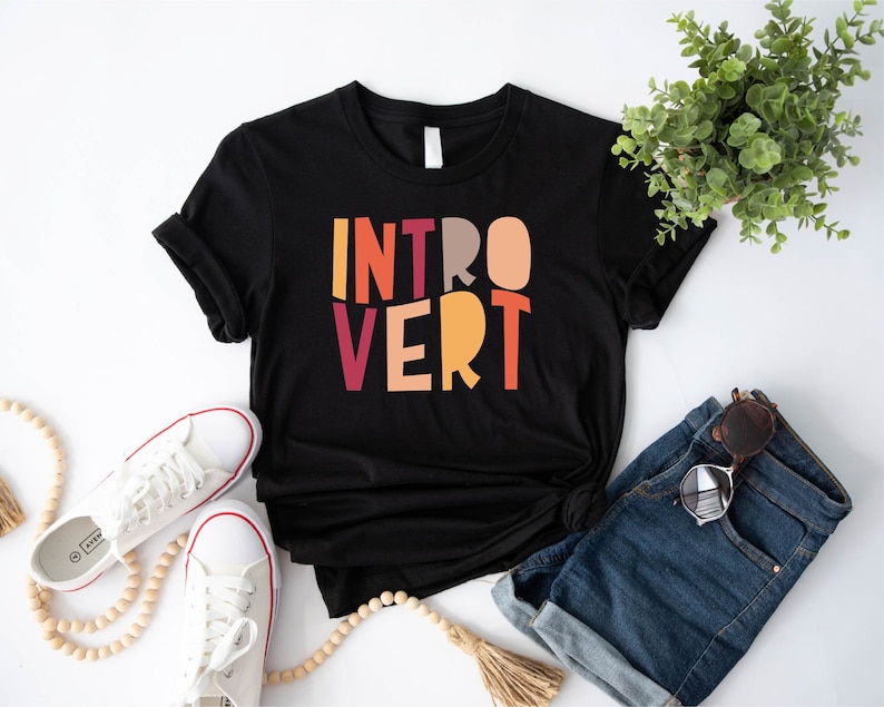 May include: Black t-shirt with the word "INTROVERT" printed in colourful block letters. The shirt is laid flat on a white surface with a pair of white trainers, a wooden bead necklace, a pair of denim shorts, and a pair of sunglasses.