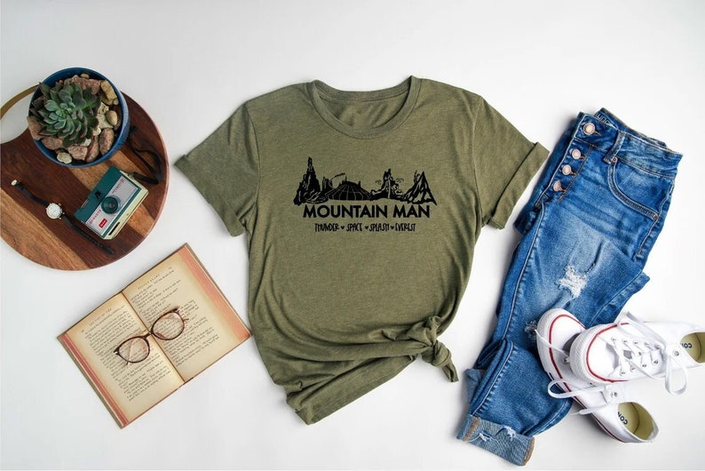 K&ouml;nnte beinhalten: Ein gr&uuml;nes T-Shirt mit einer schwarzen Bergkette und dem Text "Mountain Man" mit den Worten "Thriving - Spirit - Passion - Instinct" darunter.