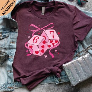 A purple t-shirt with a graphic design of two dice with the number 6 and the number 7 on them, surrounded by hearts and a pink ribbon.