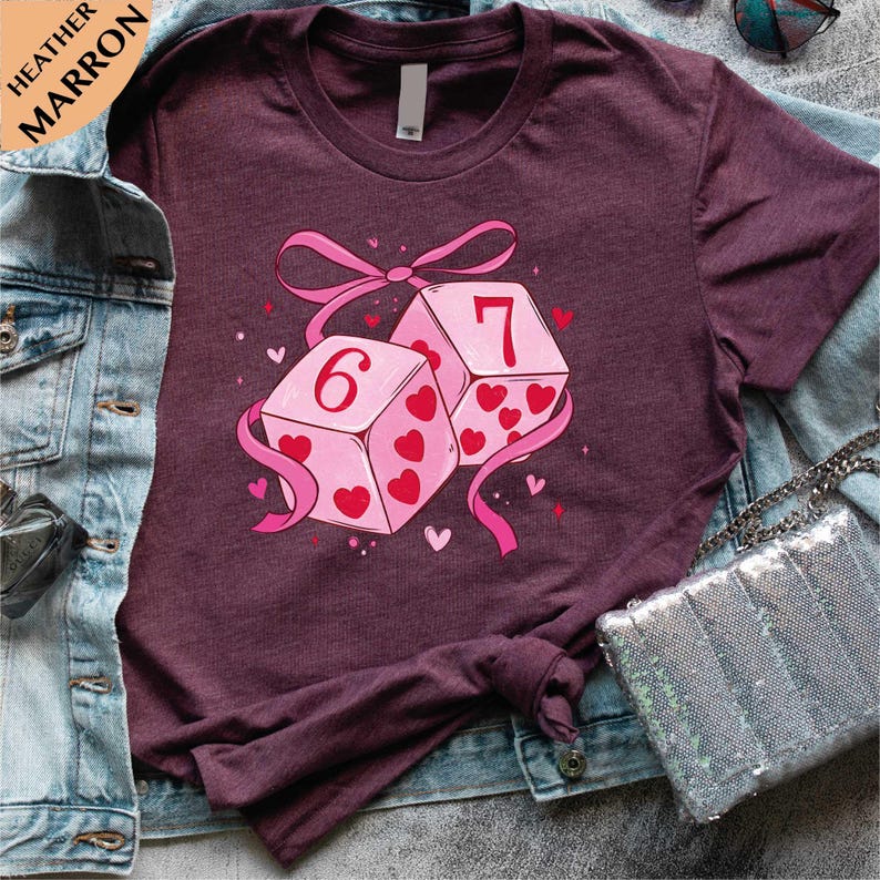 A purple t-shirt with a graphic design of two dice with the number 6 and the number 7 on them, surrounded by hearts and a pink ribbon.