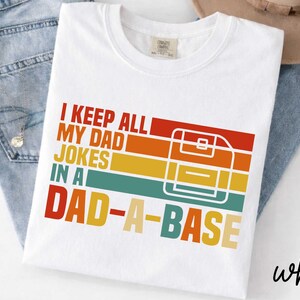May include: White t-shirt with the text "I KEEP ALL MY DAD JOKES IN A DAD-A-BASE" in a retro design. The design features a floppy disk graphic with red, orange, yellow, and teal stripes. The shirt is displayed on a pair of blue jeans.