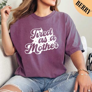 May include: A purple t-shirt with the text "Tired as a Mother" in white with a retro font and white stars.