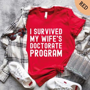 May include: A red t-shirt with the text "I SURVIVED MY WIFE'S DOCTORATE PROGRAM" in white. The shirt is paired with a plaid shirt, sunglasses, white sneakers, and a sequined purse.