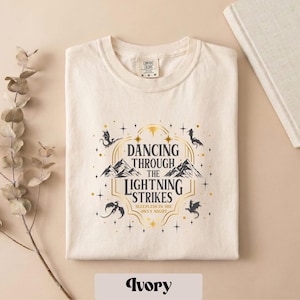 A white t-shirt with a graphic design featuring a mountain range and stars, along with the text "DANCING THROUGH THE LIGHTNING STRIKES" and "SLEEPLESS IN THE NIGHT".