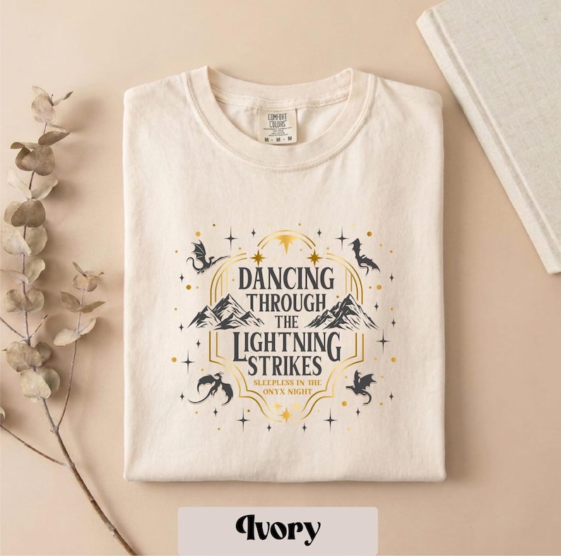 A white t-shirt with a graphic design featuring a mountain range and stars, along with the text "DANCING THROUGH THE LIGHTNING STRIKES" and "SLEEPLESS IN THE NIGHT".