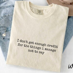 A beige t-shirt with a humorous text printed on it, placed on top of a pair of jeans.