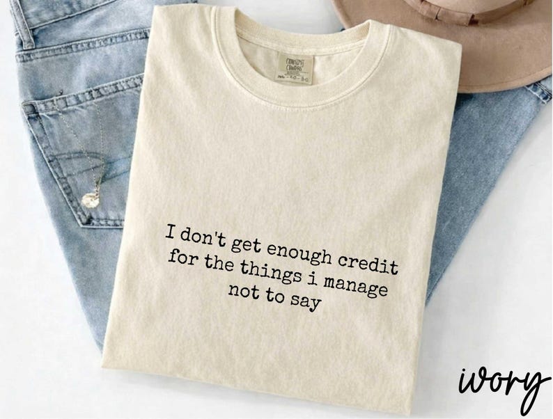 A beige t-shirt with a humorous text printed on it, placed on top of a pair of jeans.