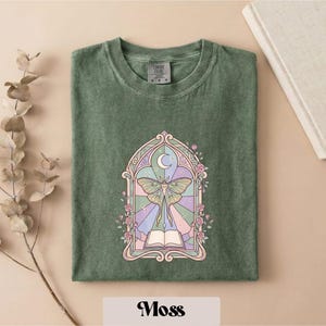 May include: A sage green t-shirt featuring a graphic print of a moth centered within an arched window frame. The design includes a crescent moon, an open book, and floral accents in pastel shades of pink, purple, and yellow.