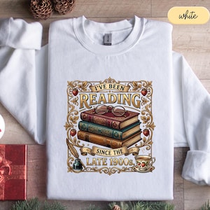 May include: A white long-sleeve sweatshirt featuring a graphic print of three stacked books, a pair of round spectacles, an inkwell with a quill, and a teacup. The text reads I'VE BEEN READING SINCE THE LATE 1900s, surrounded by a gold ornate frame.