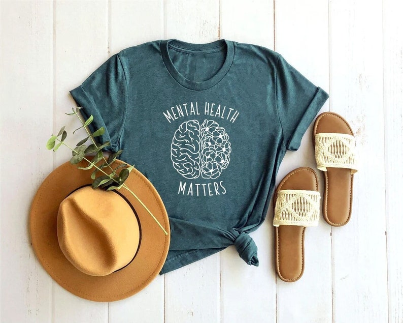 Op de afbeelding: Tealblauw T-shirt met witte tekst die "Mental Health Matters" zegt en een witte omtrek van een brein met bloemen erin.