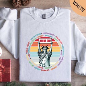 May include: White sweatshirt featuring a graphic of the Statue of Liberty holding a sign that reads "HANDS OFF!" surrounded by text. The design includes a retro sunset color scheme and various social and political terms.