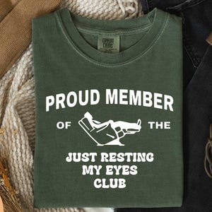 May include: A moss green t-shirt with the text "PROUD MEMBER OF THE JUST RESTING MY EYES CLUB" in white. The graphic shows a person reclining in a chair. The shirt is made of a soft material.
