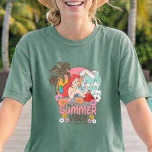 A smiling woman wearing a green t-shirt with a cartoon character and the text "Summer Vibes" is riding a bicycle.