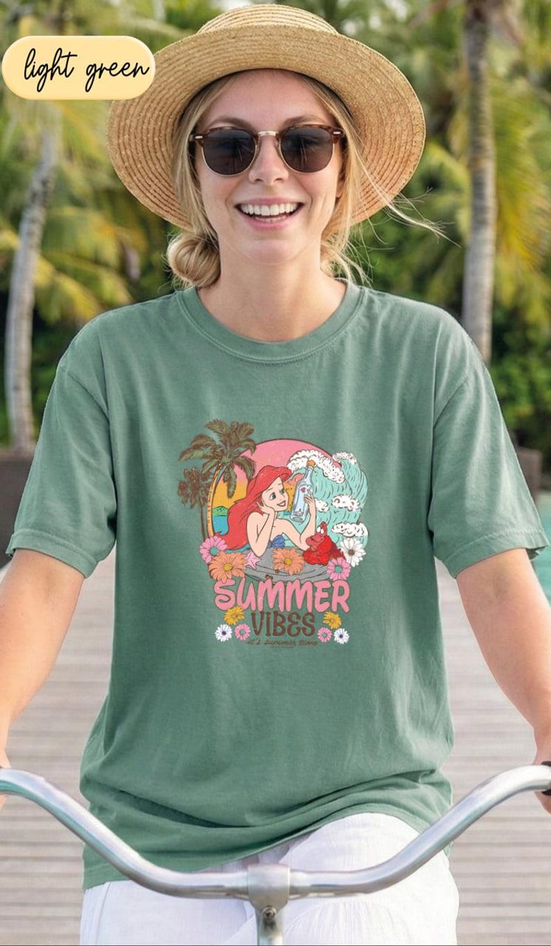 A smiling woman wearing a green t-shirt with a cartoon character and the text "Summer Vibes" is riding a bicycle.