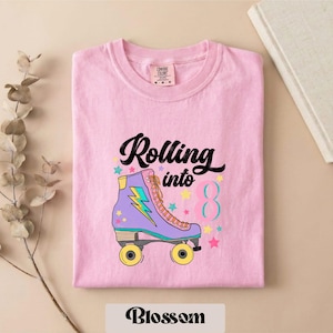 May include: A light pink t-shirt featuring a graphic of a purple roller skate with a yellow lightning bolt, surrounded by small colorful stars and the text Rolling into 8 in black and teal. The shirt has a Comfort Colors brand label.