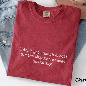 A red t-shirt with a humorous text printed on it, along with a pair of jeans and a hat, all laid out on a surface.
