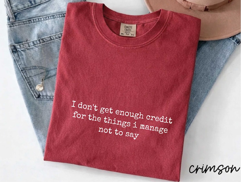 A red t-shirt with a humorous text printed on it, along with a pair of jeans and a hat, all laid out on a surface.