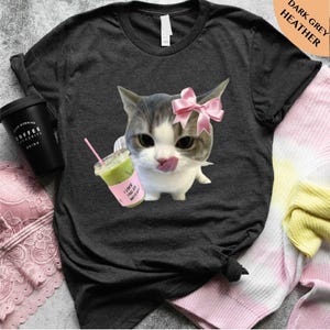 May include: Dark grey heather t-shirt featuring a cartoon cat with a pink bow, licking its lips. The cat is next to a matcha drink with a pink sleeve that says "I love you so matcha". A black coffee cup is on the left.