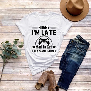 May include: White t-shirt with black text that reads "Sorry I'm Late Had To Get To A Save Point" and a black video game controller graphic.