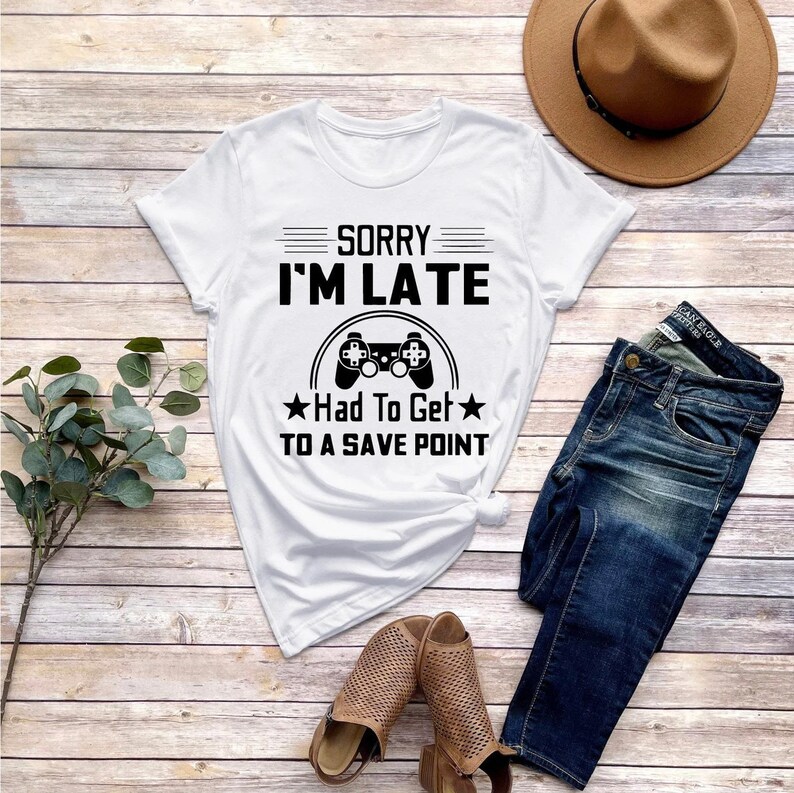 May include: White t-shirt with black text that reads "Sorry I'm Late Had To Get To A Save Point" and a black video game controller graphic.