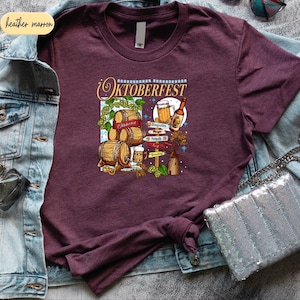 May include: A maroon short-sleeve t-shirt featuring a graphic print with the text Oktoberfest. The design includes illustrations of wooden beer barrels, a beer mug, a glass bottle, a windmill, and hop vines, all arranged in a colorful composition.