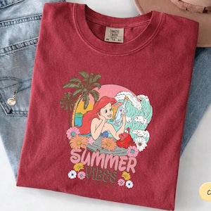 A red t-shirt with a colorful graphic design depicting a mermaid surrounded by tropical elements, including a palm tree and flowers, along with the text "Summer Vibes".