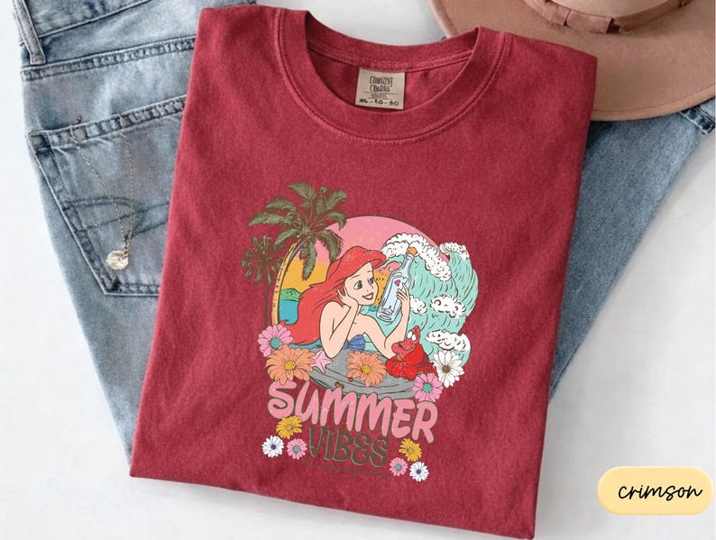 A red t-shirt with a colorful graphic design depicting a mermaid surrounded by tropical elements, including a palm tree and flowers, along with the text "Summer Vibes".