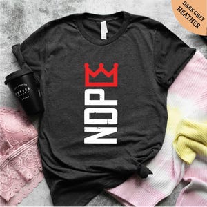 Pu&ograve; includere: T-shirt grigio scuro m&eacute;lange con la scritta "NDPE" in lettere bianche verticali e una grafica a corona rossa. Nella foto sono visibili una tazza di caff&egrave; nera e un capo in pizzo rosa.