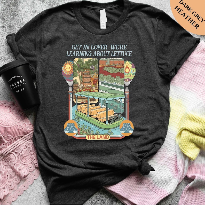 a black t-shirt with a colorful graphic design depicting a boat and a tropical landscape, along with the text "GET IN LOSER WE'RE LEARNING ABOUT LACTUCE" and "THE LAND".