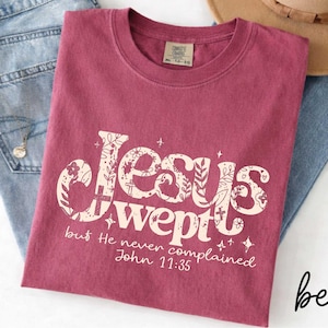May include: Berry-colored t-shirt with the text "Jesus Wept" in a decorative font, with floral and cross accents. The text "but He never complained John 11:35" is below. The shirt is folded on top of blue jeans and a brown hat.
