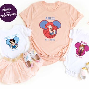 Disney Princess-themed clothing and accessories, including a pink t-shirt with the character Ariel, a white t-shirt with the character Snow White, and a pink onesie with the character Anna.