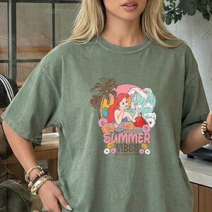 A woman wearing a green t-shirt with a graphic design of a mermaid and the text "Summer Vibes" is standing in front of a window.