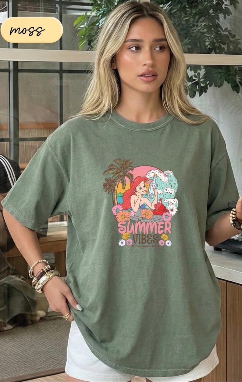 A woman wearing a green t-shirt with a graphic design of a mermaid and the text "Summer Vibes" is standing in front of a window.