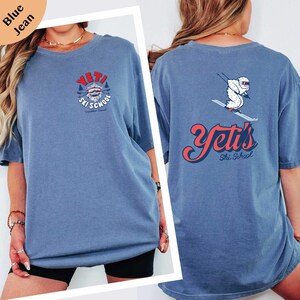 A woman wearing a blue t-shirt with a graphic design on the back, including a skiing-related image and text.