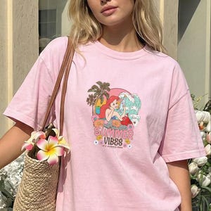 A young woman wearing a pink t-shirt with a cartoon mermaid graphic and the text "Summer Vibes" is standing in front of a building. She is holding a woven straw bag with a flower attached to it.