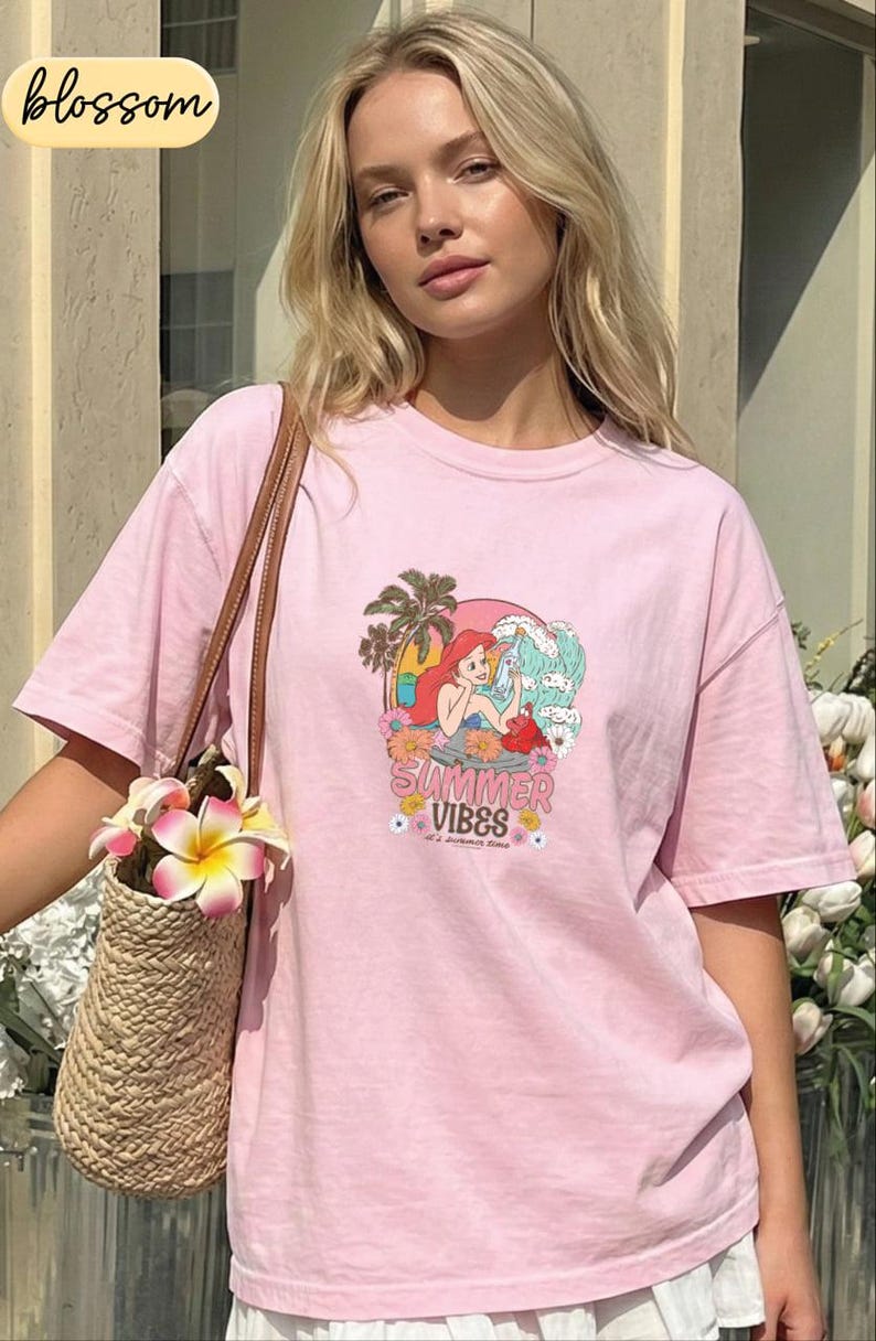 A young woman wearing a pink t-shirt with a cartoon mermaid graphic and the text "Summer Vibes" is standing in front of a building. She is holding a woven straw bag with a flower attached to it.