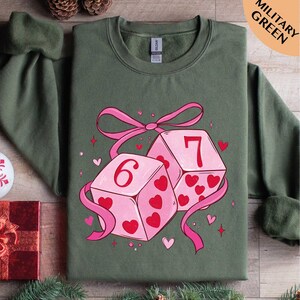 A green t-shirt with a graphic design featuring two pink dice with the number 6 and 7 on them, surrounded by hearts and ribbons.