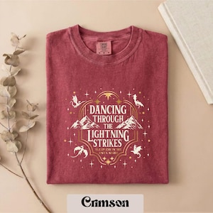 A red t-shirt with a white and gold design featuring the text "DANCING THROUGH THE LIGHTNING STRIKES ONLY NIGHT" and a dragon silhouette. The t-shirt is folded and placed on a beige background, with a plant branch and a book visible in the background.