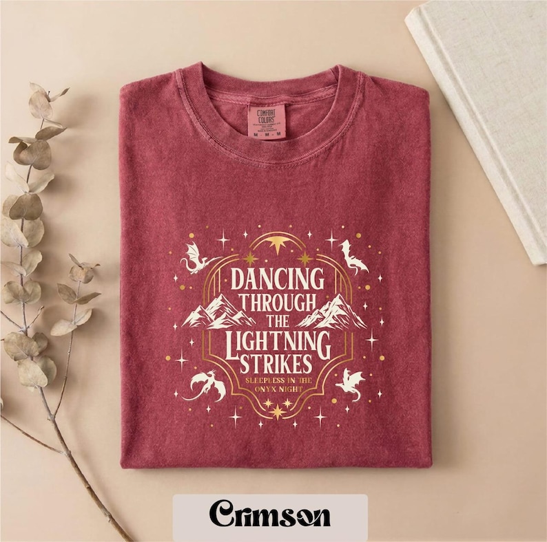 A red t-shirt with a white and gold design featuring the text "DANCING THROUGH THE LIGHTNING STRIKES ONLY NIGHT" and a dragon silhouette. The t-shirt is folded and placed on a beige background, with a plant branch and a book visible in the background.
