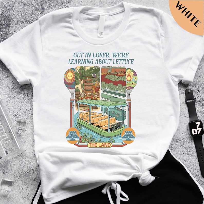 A white t-shirt with a colorful graphic design depicting a boat and various elements related to the phrase "Get in Loser, Were Learning about Lettuce" printed on it.