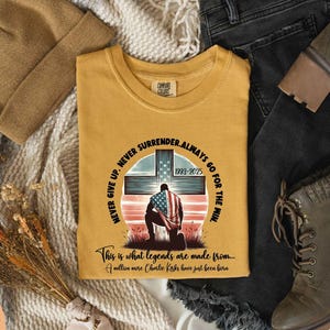 May include: Mustard-colored t-shirt with a graphic of a kneeling figure draped in an American flag in front of a cross. Text includes "NEVER GIVE UP. NEVER SURRENDER. ALWAYS GO FOR THE WIN" and "This is what legends are made from..."
