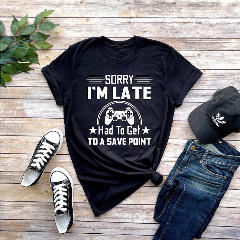 May include: Black t-shirt with white text that reads "Sorry I'm Late Had To Get To A Save Point" with a white video game controller graphic.