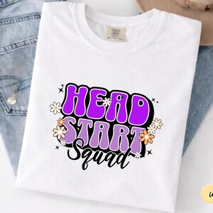 May include: A white short-sleeved t-shirt featuring a graphic print with the text HEAD START SQUAD in bold purple and light purple bubble letters. The design is accented with small white and orange daisy flowers and black starburst icons.