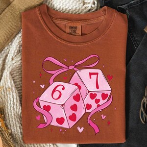 a brown t-shirt with a graphic design of two pink dice with the number 6 on them, surrounded by hearts and ribbons.