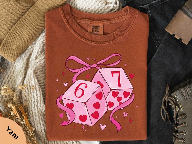 a brown t-shirt with a graphic design of two pink dice with the number 6 on them, surrounded by hearts and ribbons.