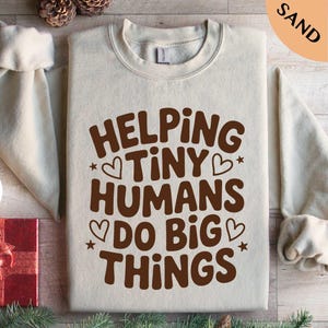 May include: A sand-colored sweatshirt with the phrase "HELPING TINY HUMANS DO BIG THINGS" in brown lettering. The design includes hearts and stars. The sweatshirt has long sleeves and a crew neck.