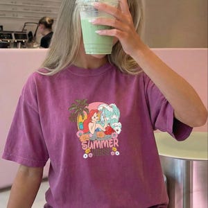 A person wearing a purple t-shirt with a graphic of a mermaid and the text "Summer vibes" is holding a green drink up to their face.