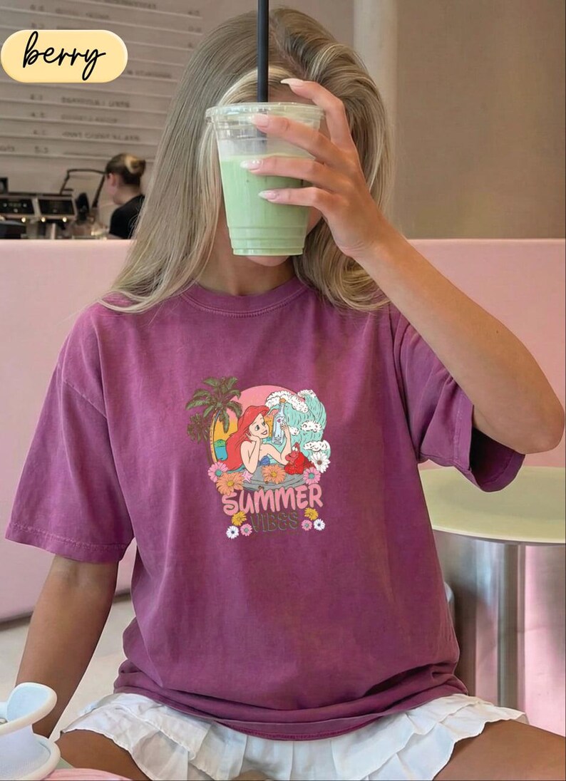 A person wearing a purple t-shirt with a graphic of a mermaid and the text "Summer vibes" is holding a green drink up to their face.