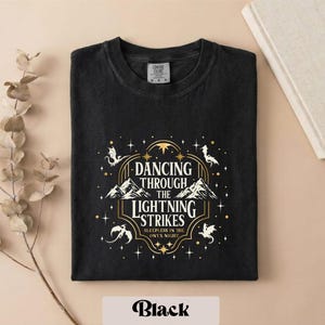 A black t-shirt with a graphic design featuring a mountain range and stars, along with the text "DANCING THROUGH THE LIGHTNING STRIKES" and "SLEEPY NIGHT".